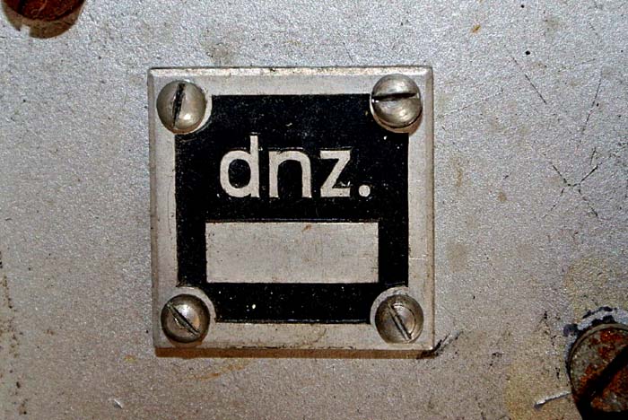 Tankový přijímač Ukw.E.e - čtvercový štítek s plastickým krycím kódem "dnz." patřícím firmě Schwarzwälder Apparate-Bauanstalt, August Schwer Söhne, Villingen/Schwarzwald. Štítek je umístěný skrytě na chassis přístroje pod předním panelem.